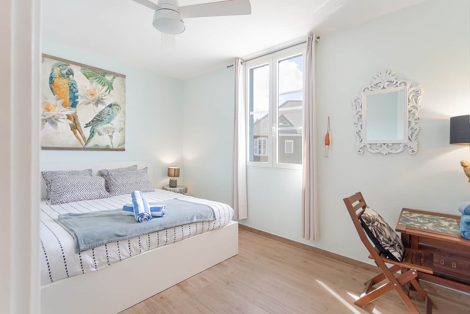 A bedroom with a bed, a desk, a chair, a ceiling fan, a window, and a painting of birds.