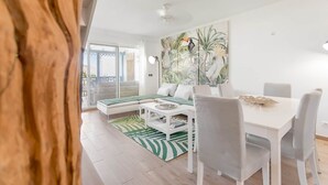 A modern living room with a white sofa, a dining table with chairs, and a large tropical-themed painting on the wall.