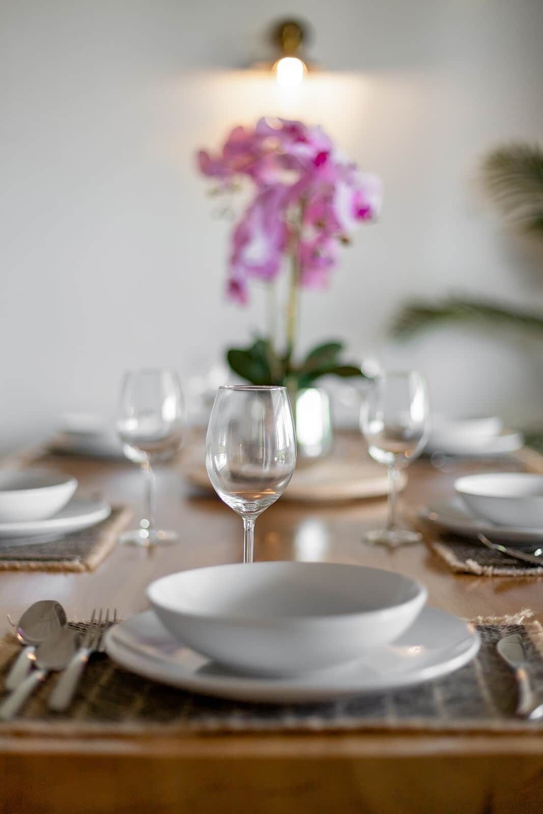 Een eettafel gedekt met witte borden, heldere glazen en bestek, met een vaas roze bloemen op de achtergrond.