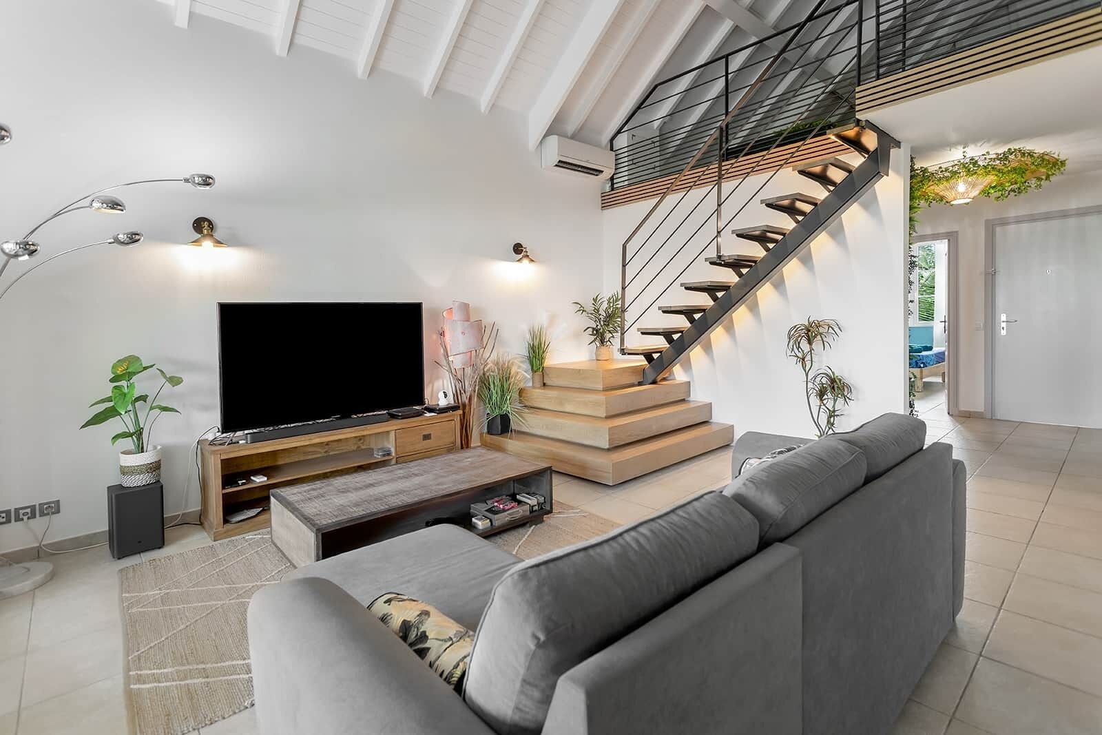Un salon moderne doté d’un grand téléviseur à écran plat, d’un canapé gris, d’un meuble de rangement en bois et d’un escalier avec une rampe en verre.