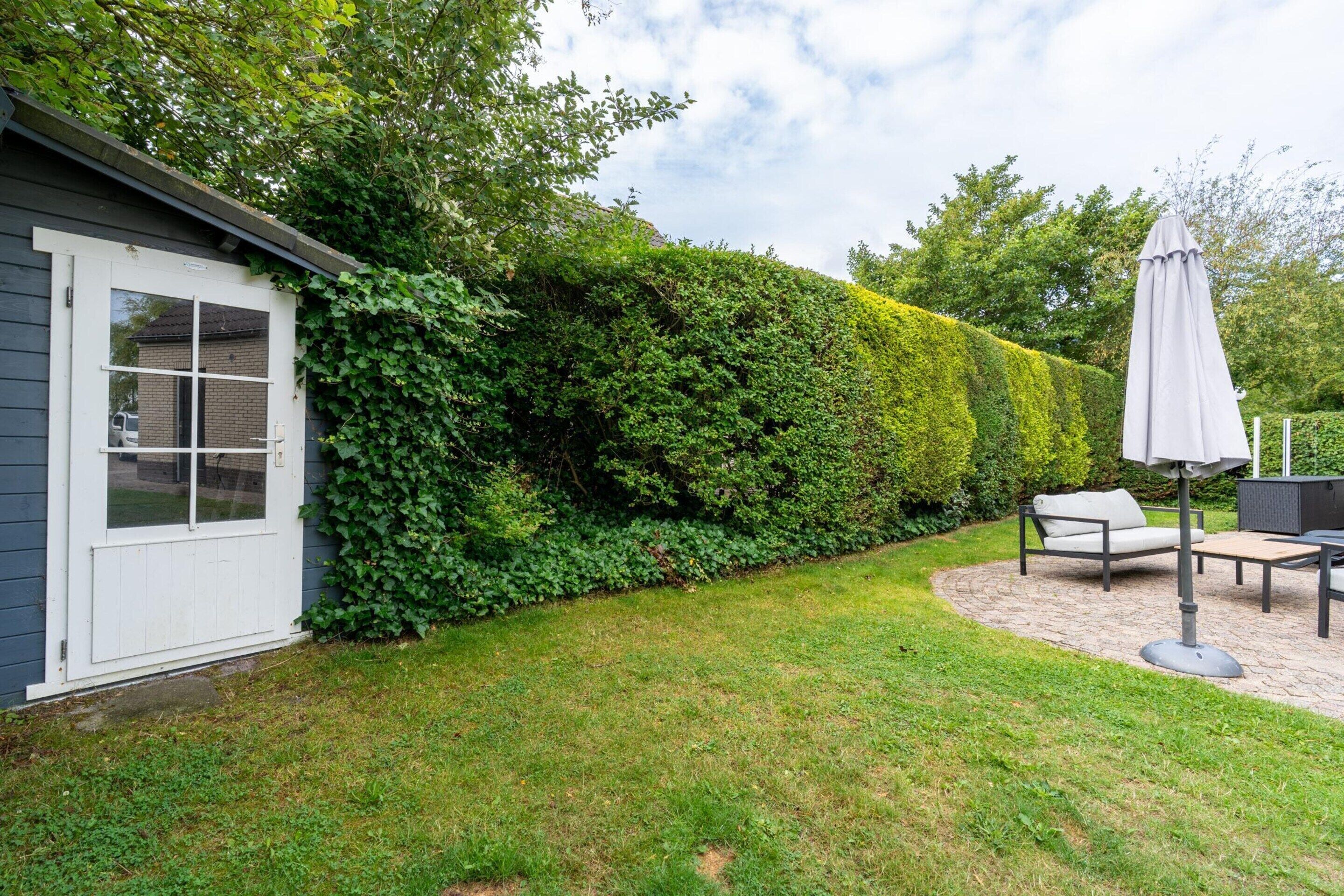 Ein gepflegter Garten mit einer Hecke, einer Terrasse mit Gartenmöbeln und ein Haus mit einer weißen Tür.