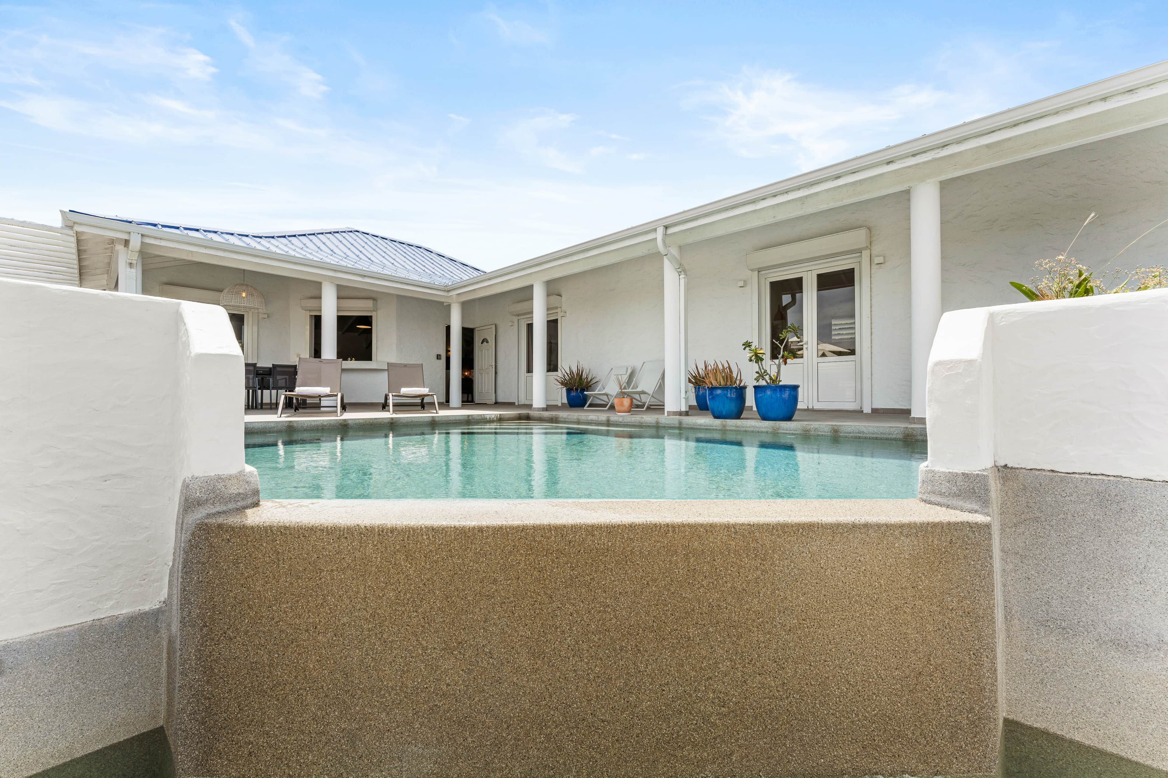 A pool area with a clear blue pool, white columns, and a covered seating area with chairs and tables.