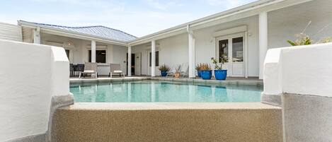 A pool area with a clear blue pool, white columns, and a covered seating area with chairs and tables.