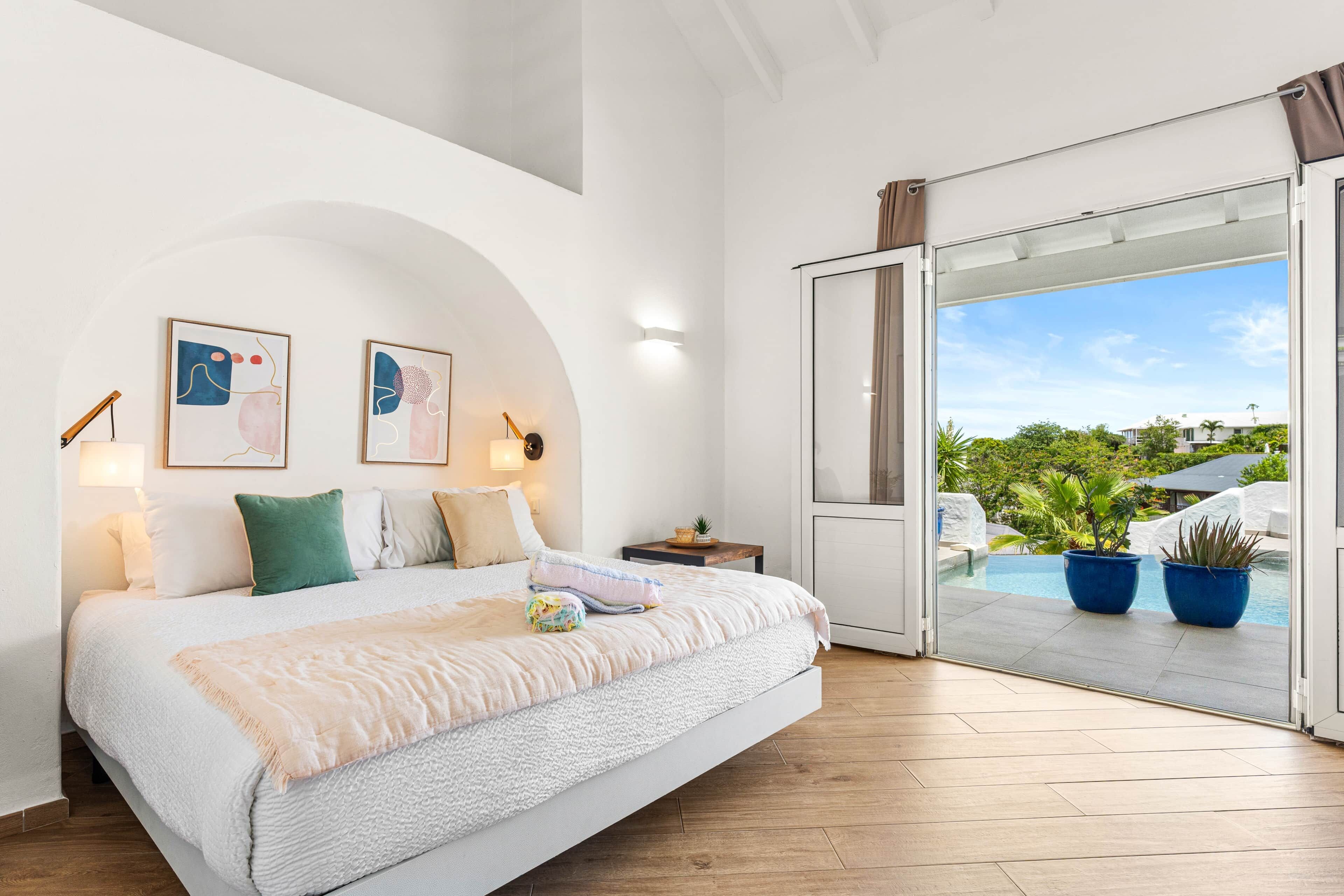 A bedroom with a bed, pillows, a nightstand, wall art, and a view of an outdoor pool area.