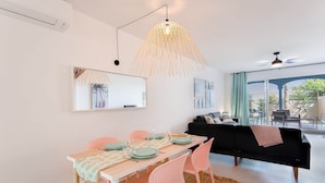 A modern living room with a dining area, featuring a black sofa, pink chairs, a wooden dining table, and a large window with a view of the outdoors.
