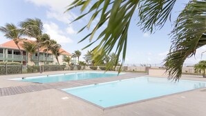 Une piscine rectangulaire avec une terrasse en bois, entourée de palmiers et d’une clôture.