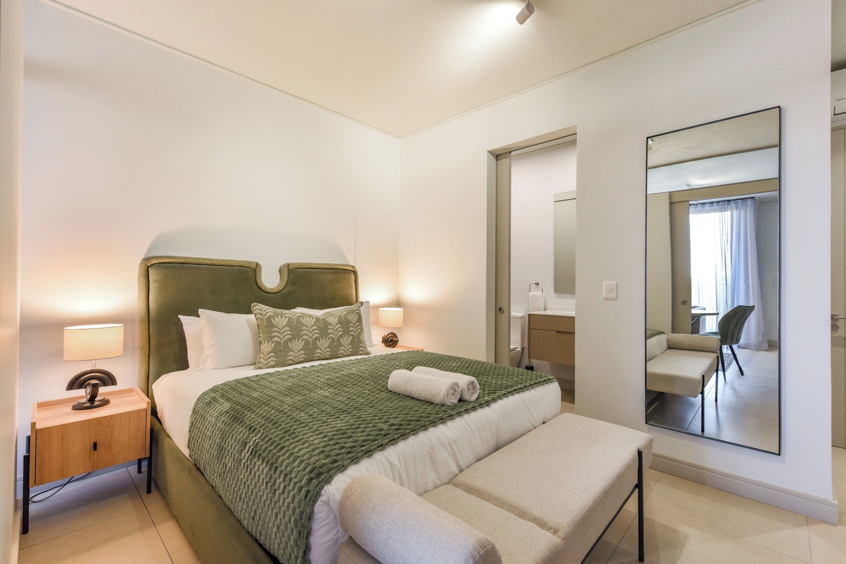 Habitación de hotel moderna con una cama grande, una mesita de noche con lámpara, un espejo y vistas a un balcón.
