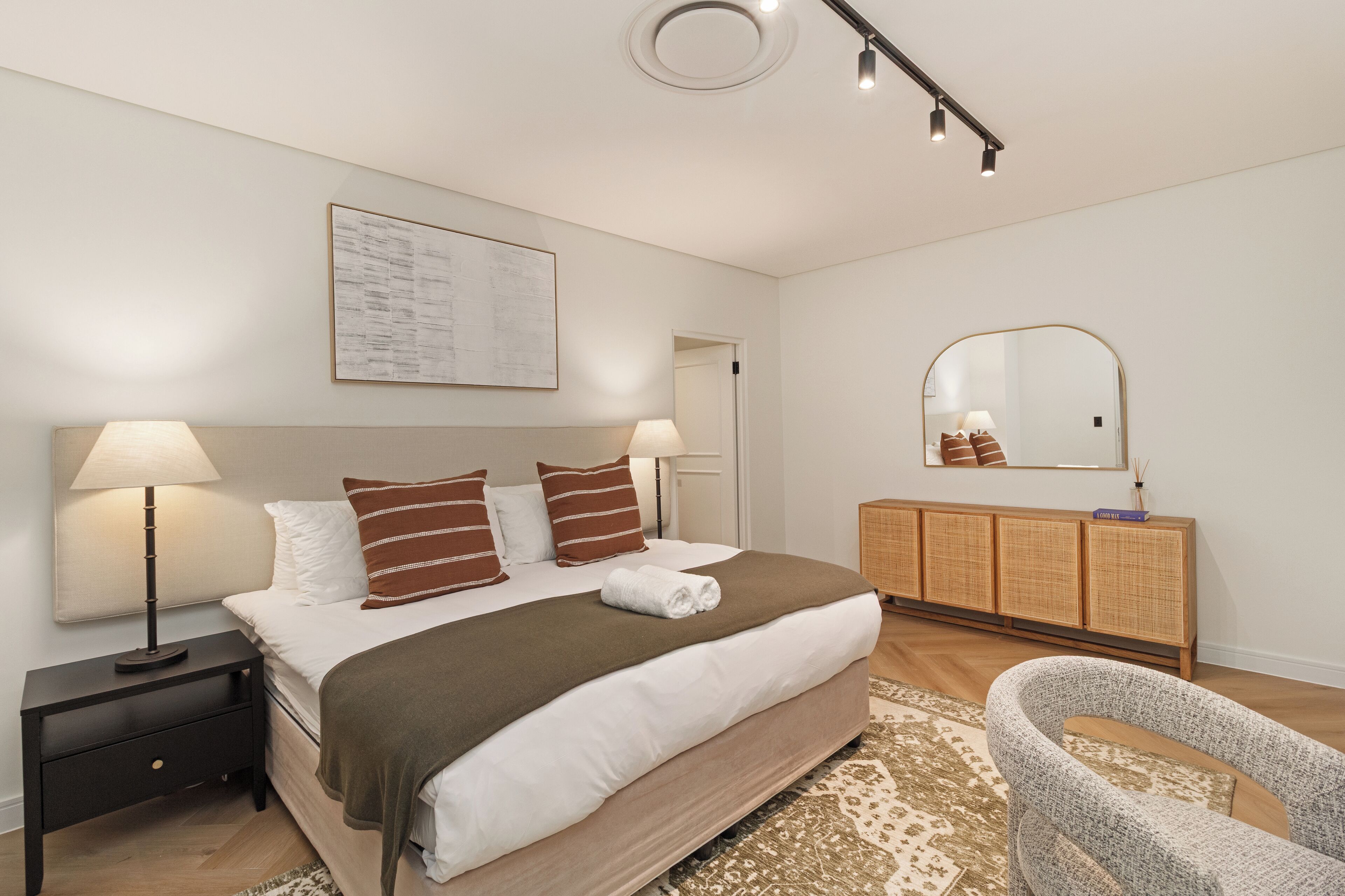 A modern bedroom with a large bed, bedside lamps, a wooden cabinet, and a mirror.