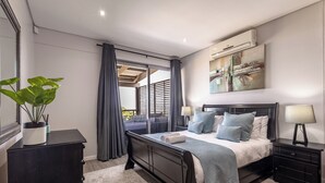 A modern bedroom with a large bed, a nightstand, a dresser, a potted plant, and a view of the outdoors.