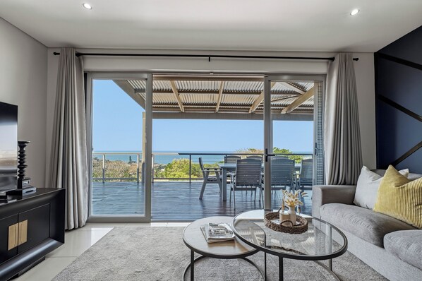 A modern living room with a large sliding glass door leading to a balcony with a view of the sea.