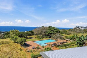 Una piscina con vista al océano, rodeada de vegetación y un área cercada con sillones de descanso.