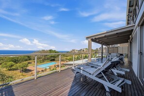 A wooden deck with lounge chairs and a view of a swimming pool and ocean.