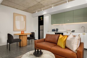 Une cuisine moderne avec des meubles de couleur verte, un coin repas doté d’une table ronde en bois, et un canapé en cuir marron agrémenté de coussins jaunes et blancs.