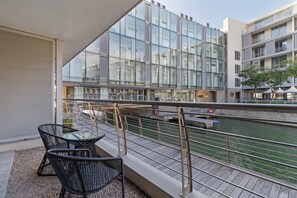 Un edifici modern amb grans finestres de vidre, un balcó amb mobiliari exterior i vista al riu.