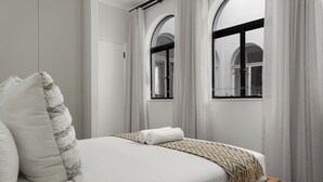 A neatly made bed with a white comforter and a patterned throw blanket, a wooden bench at the foot of the bed, and a view of a building through a window with a black frame. - Venice-Inspired Penthouse with City Views (Cape Town)