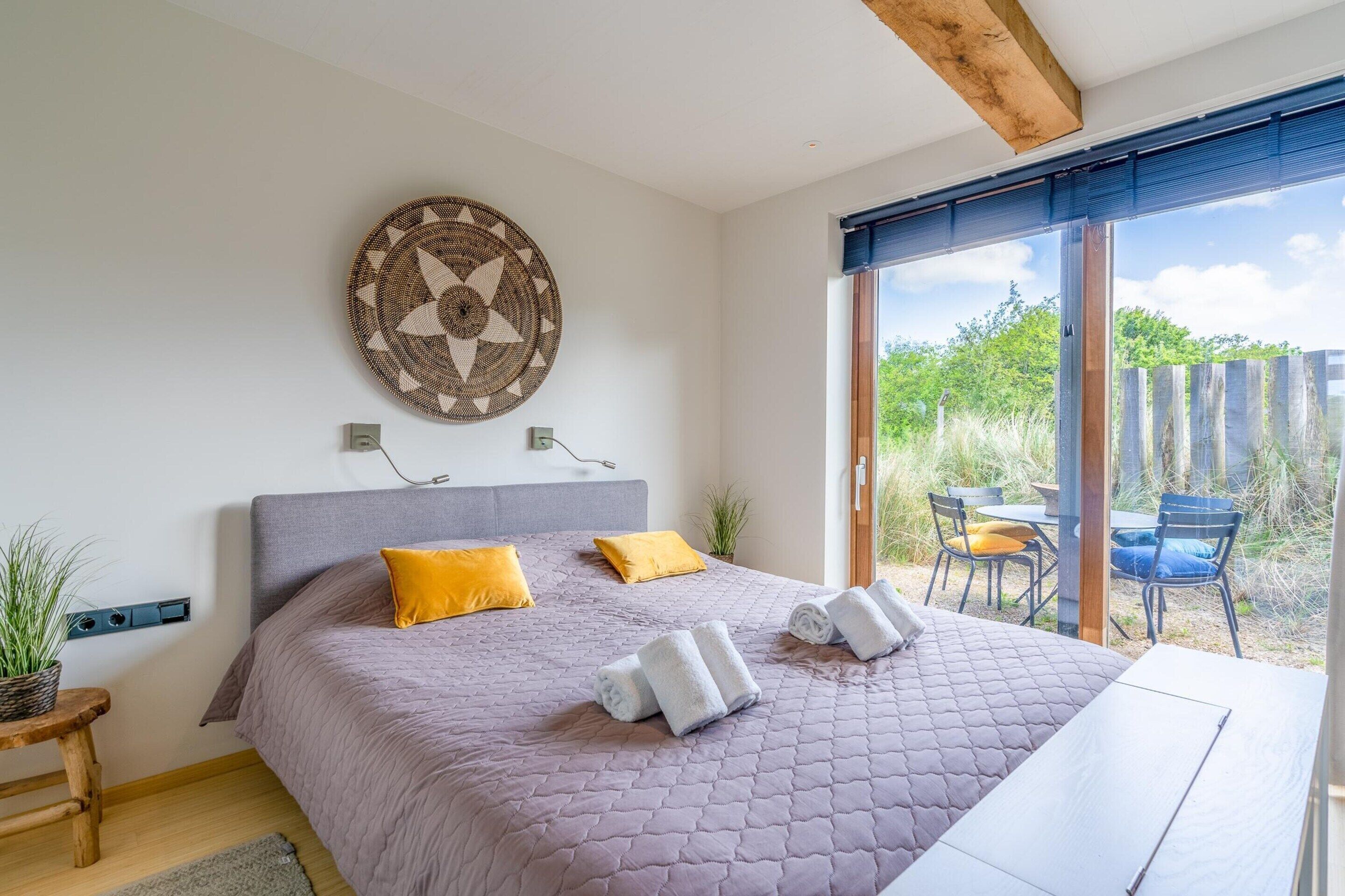 A bedroom with a large bed, two pillows, a wall art piece, and a view of the outside.