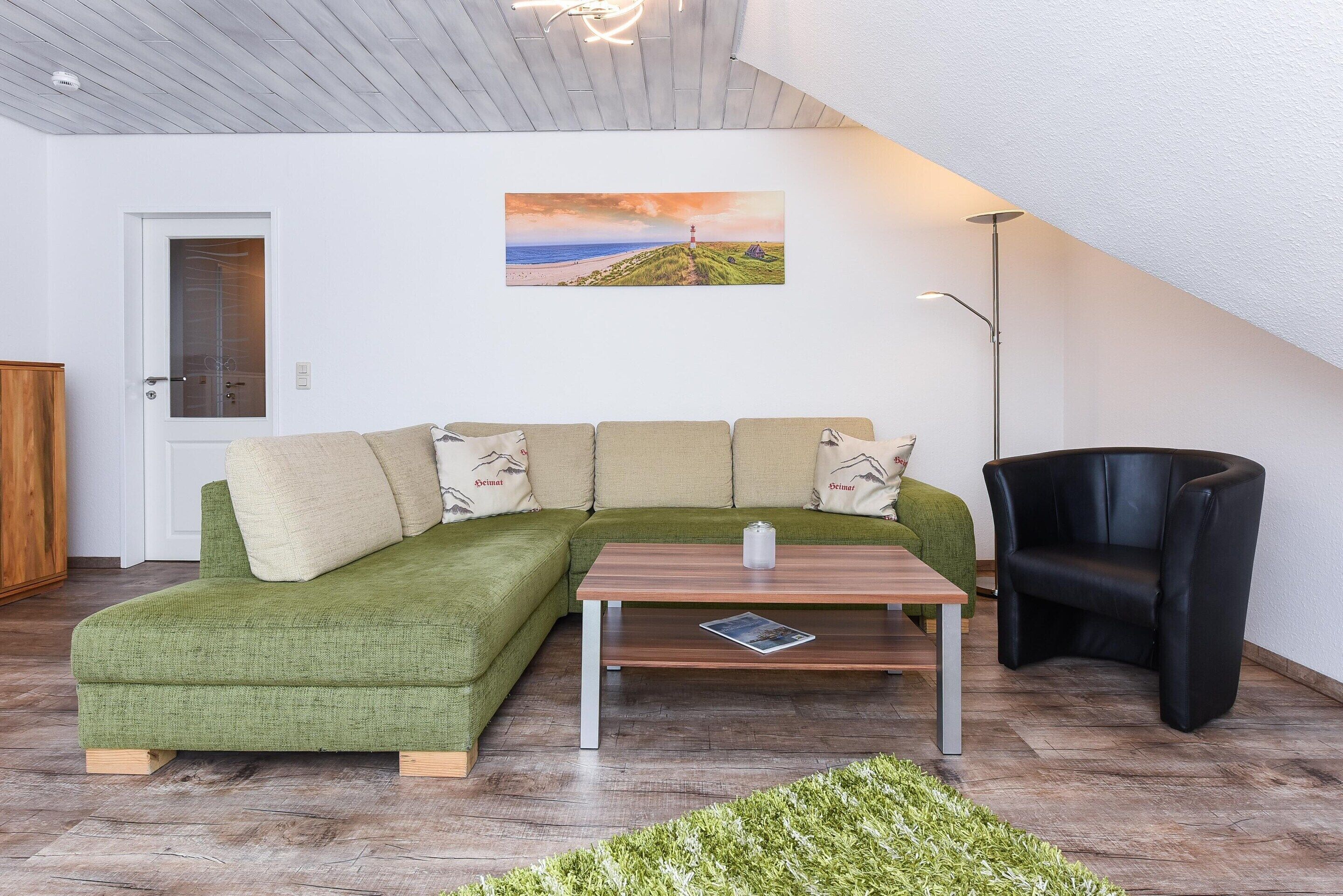 A modern living room with a green sectional sofa, a wooden coffee table, a black armchair, and a painting of a coastal landscape on the wall.