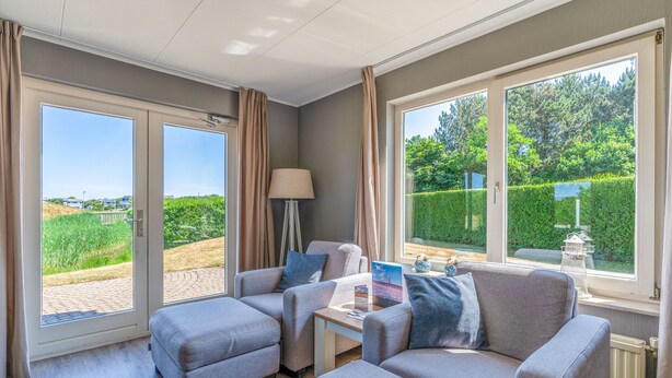 Een moderne woonkamer met grijze banken, een salontafel en een lamp. Grote ramen bieden uitzicht op groen en een verafgelegen landschap.