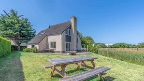 Een huis met een schuin dak, schoorsteen en een grote houten picknicktafel in de voortuin.