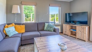 Een moderne woonkamer met een grijze bank, een houten salontafel, een televisie op een houten meubel en twee ramen met zonwering.