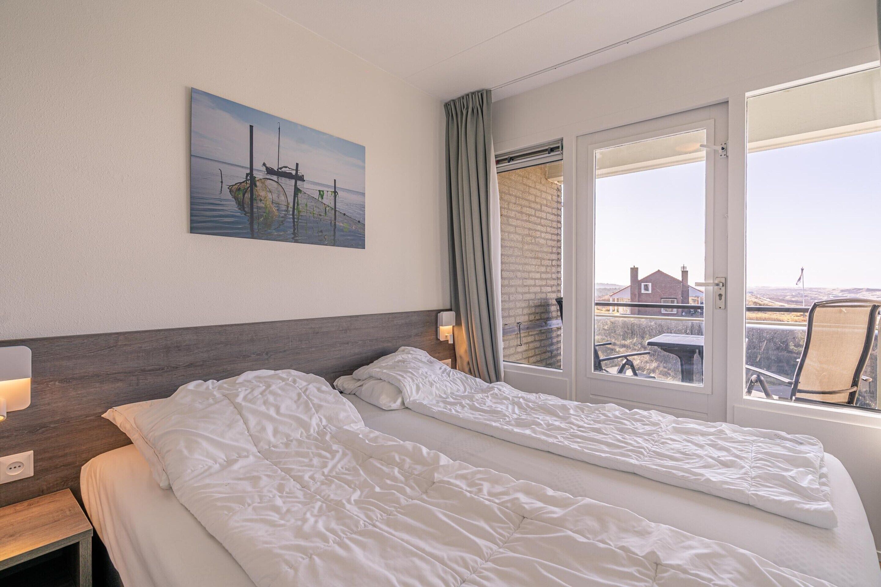 Ein Hotelzimmer mit zwei Betten, einem Bild an der Wand, einem Fenster mit Vorhängen und Blick nach draußen.