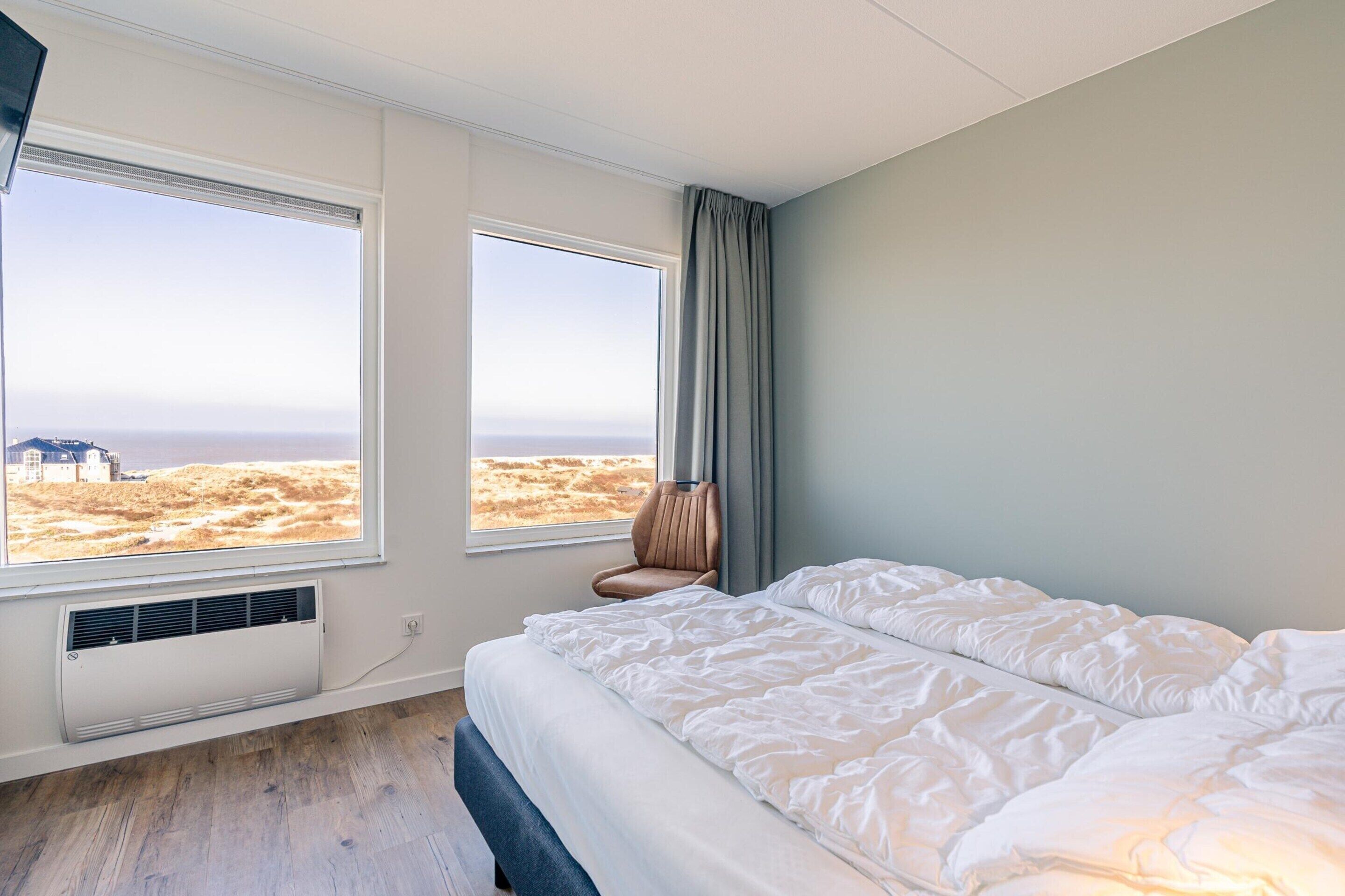 A bedroom with a large bed, two windows with a view of a beach, and a chair near the windows.
