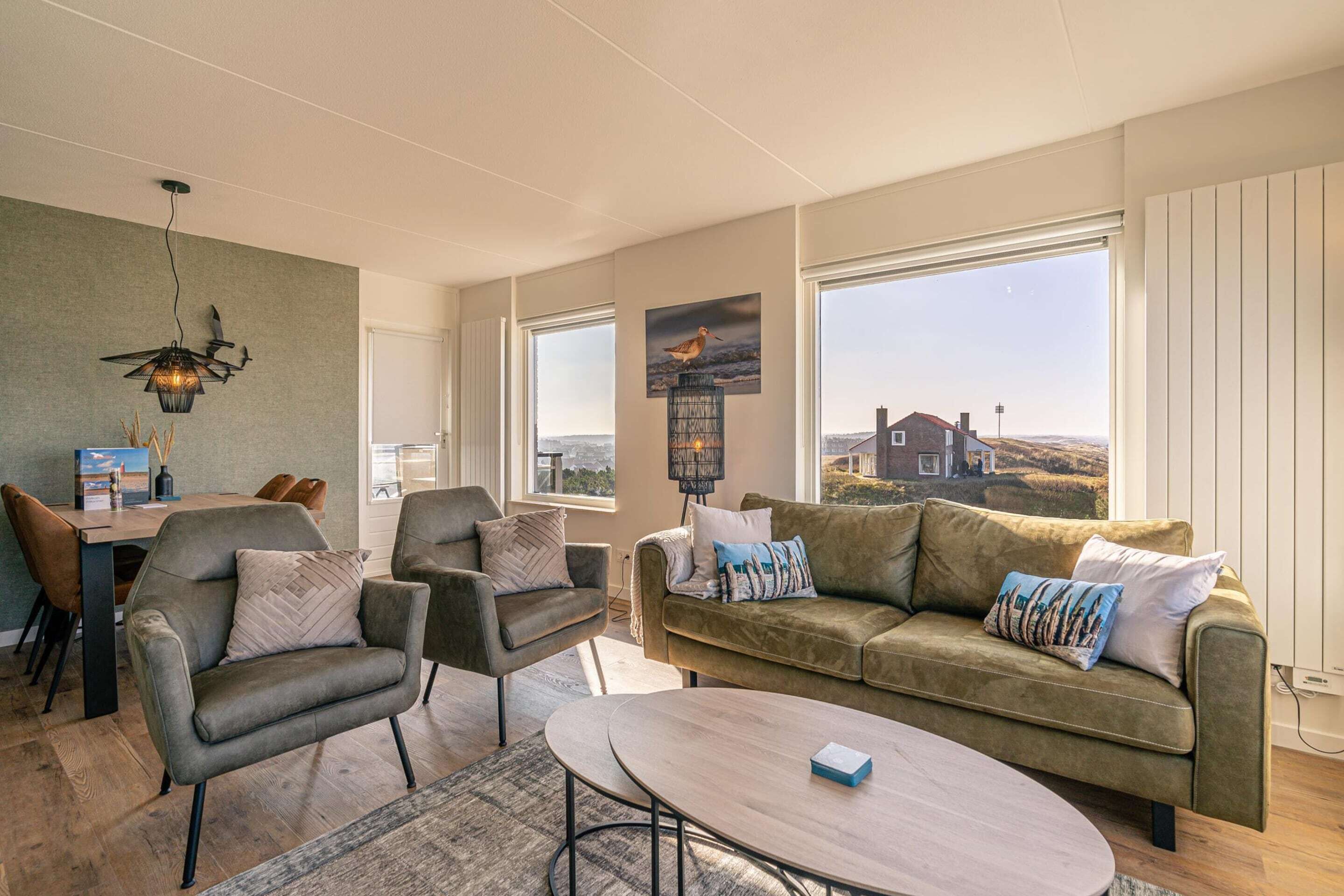 A modern living room with a sofa, chairs, a coffee table, and large windows offering a view of the countryside.
