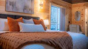 A cozy bedroom with a large bed, a nightstand, a lamp, and a window with a view of trees.