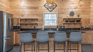 A modern kitchen with wooden walls, a stainless steel refrigerator, a microwave, a sink, and bar stools.
