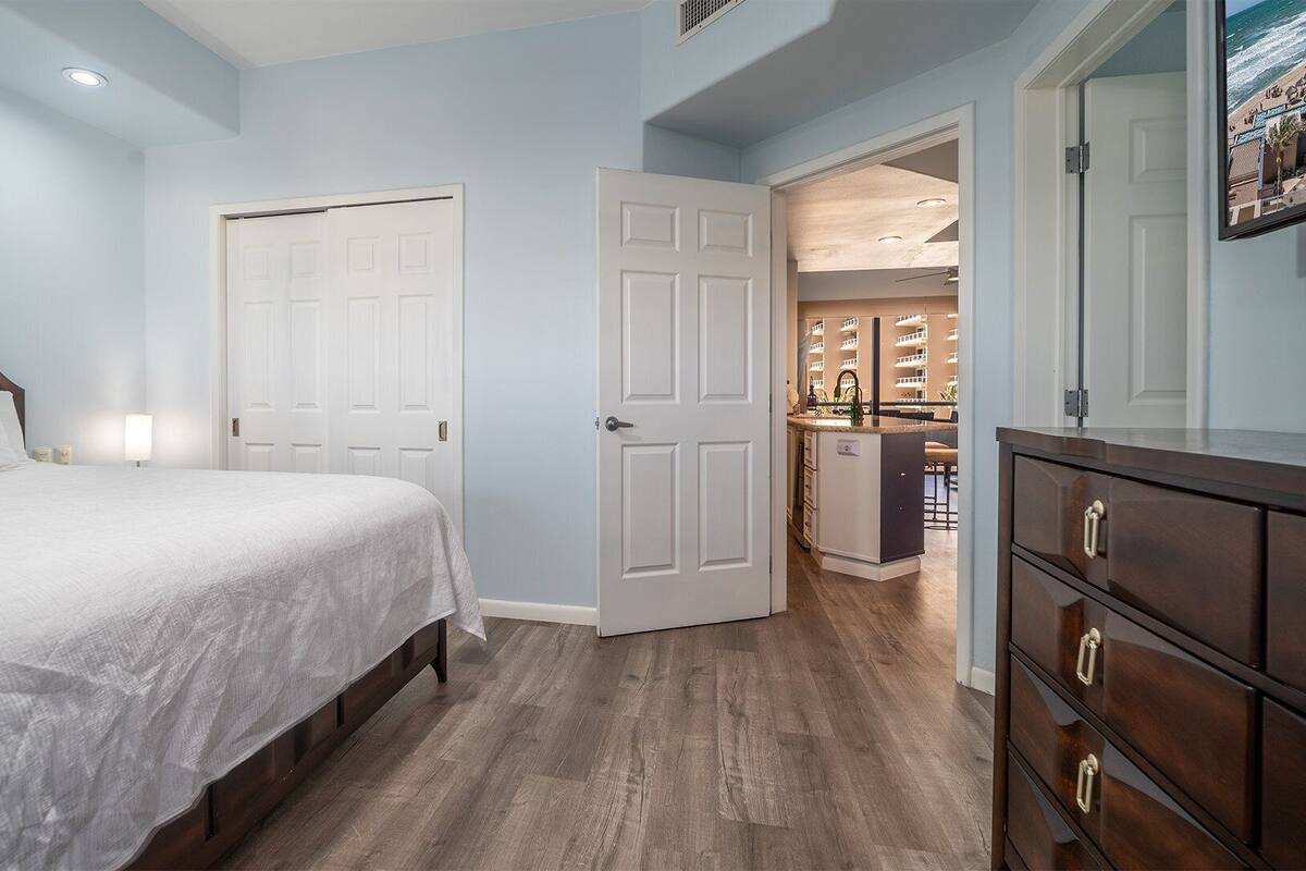 A bedroom with a bed, a dresser, and a door leading to a bathroom and kitchen area.