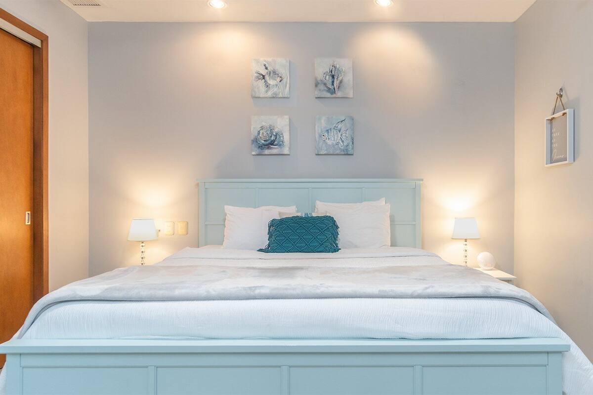 A bedroom with a light blue bed, white bedding, and a teal throw pillow. There are four wall-mounted artworks above the bed, and a framed quote on the wall to the right.