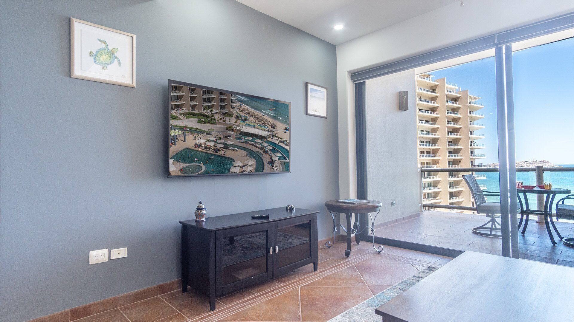 A modern living room with a large painting of a coastal town, a dark wooden cabinet, and a view of a balcony with outdoor furniture.