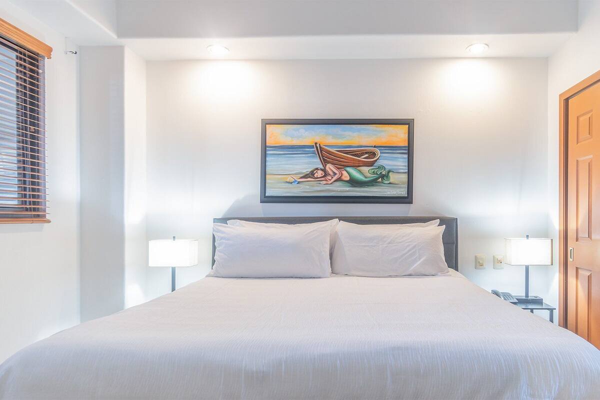 A bedroom with a bed, two bedside lamps, a painting on the wall, and a wooden door.