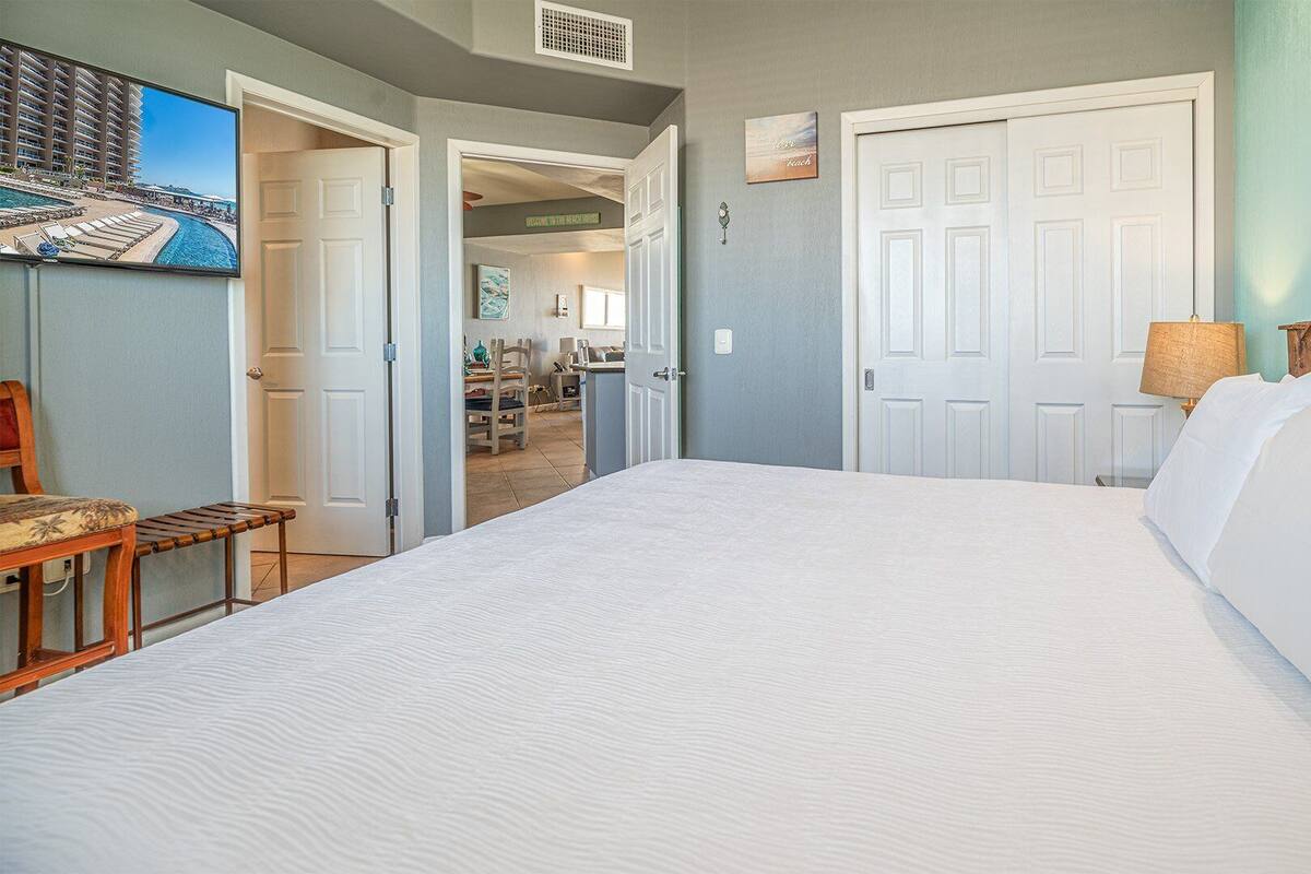A bedroom with a large bed, a wooden headboard, a nightstand with a lamp, and a view of a beach through an open door.