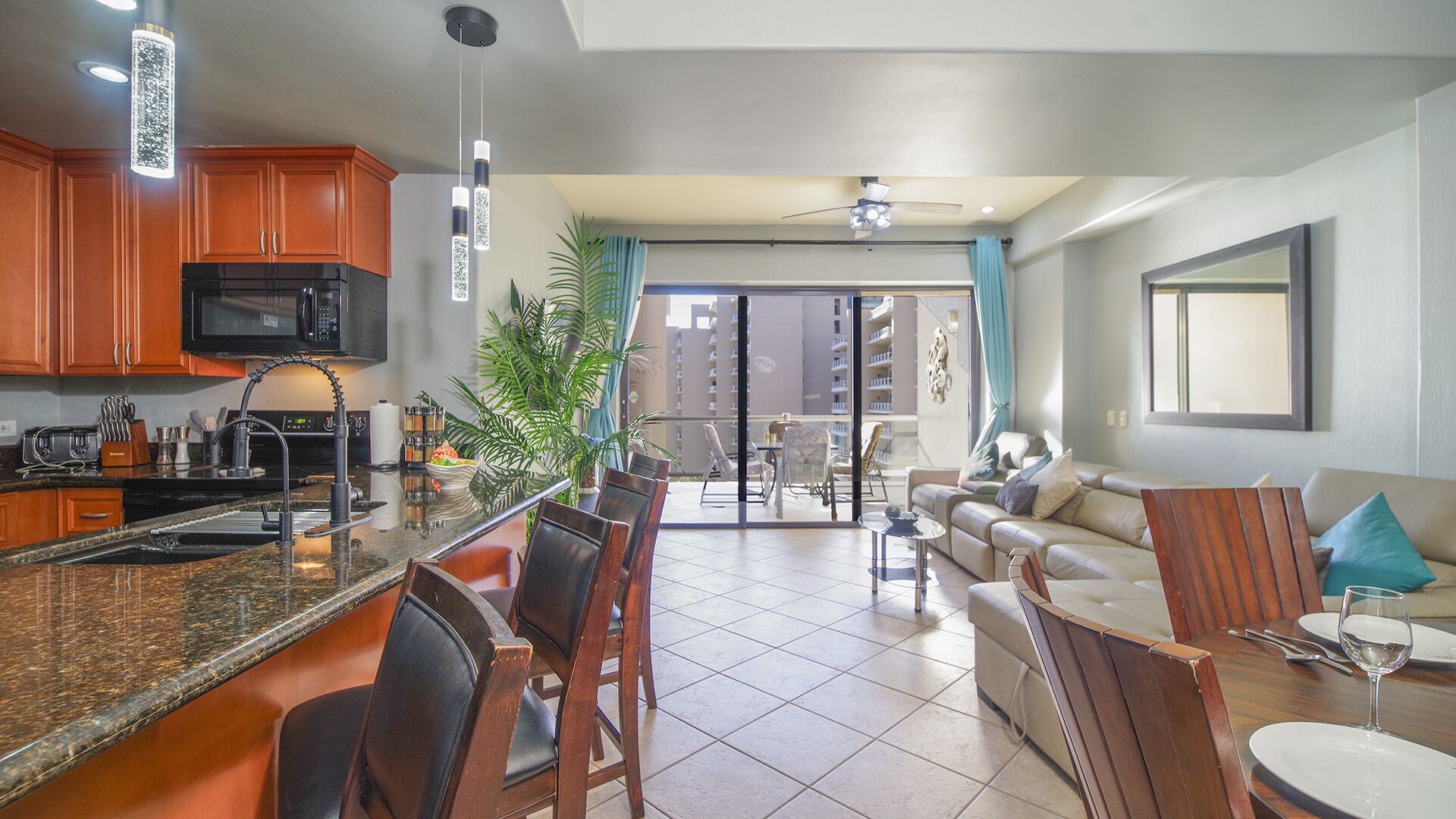 A modern kitchen with granite countertops, wooden cabinets, and a black microwave. There is a dining area with a wooden table and chairs, and a living room with a sofa and a coffee table.