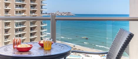 Ein Balkon mit Tisch für zwei Personen, Blick auf Strand und ferne Stadtlandschaft.