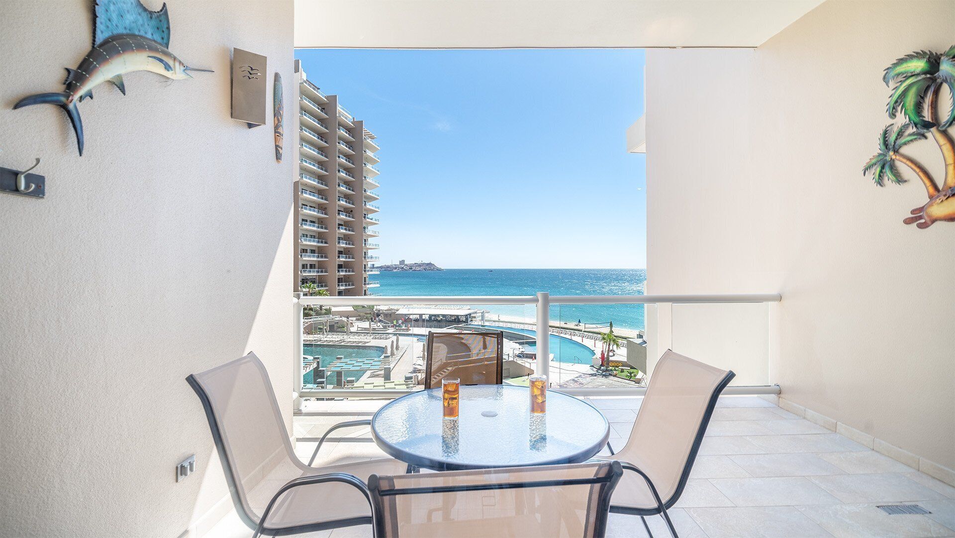 A balcony with a table and chairs, overlooking a pool and a beach with a high-rise building in the background.
