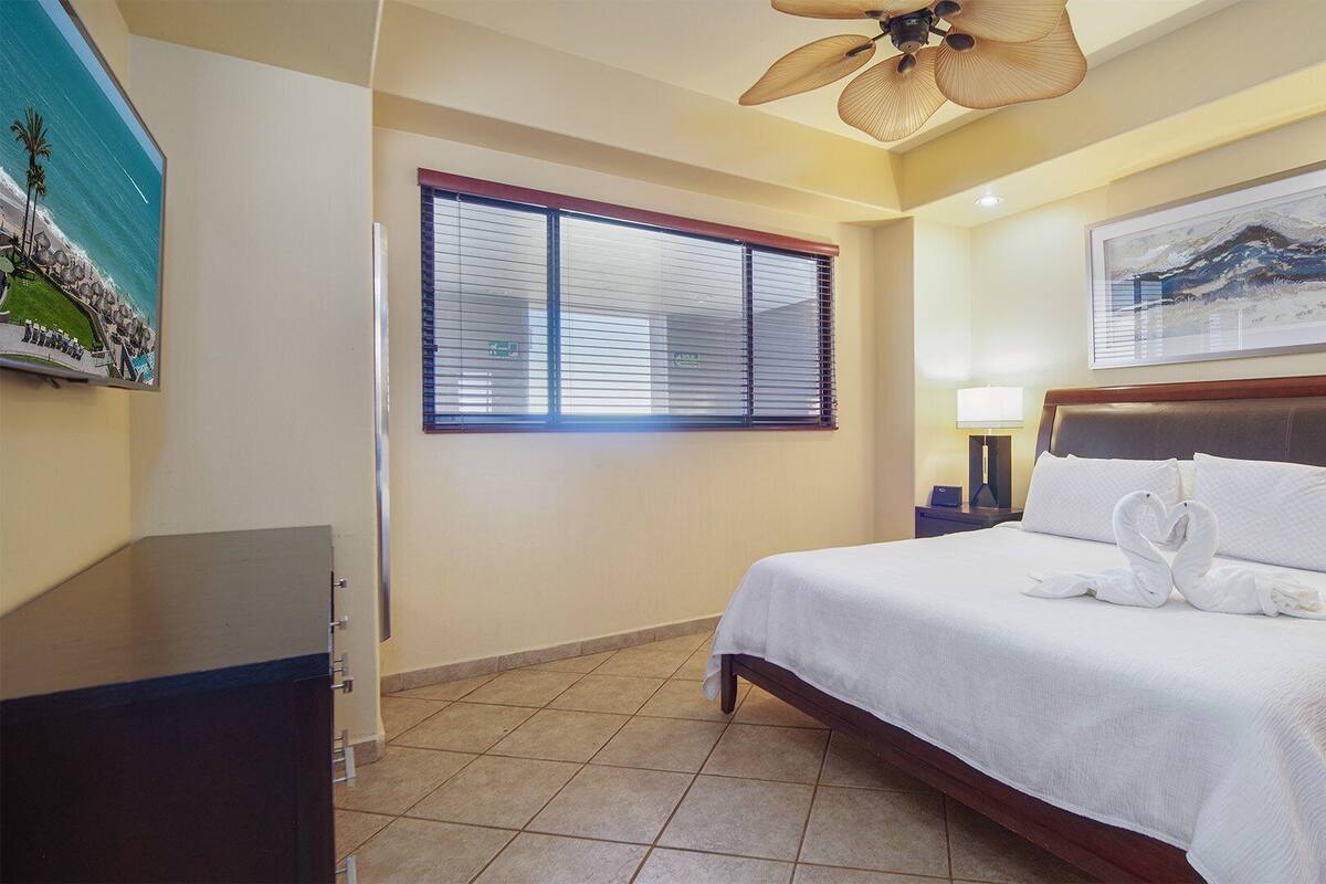 A hotel room with a bed, a ceiling fan, a window with blinds, a painting on the wall, and a nightstand with a lamp.