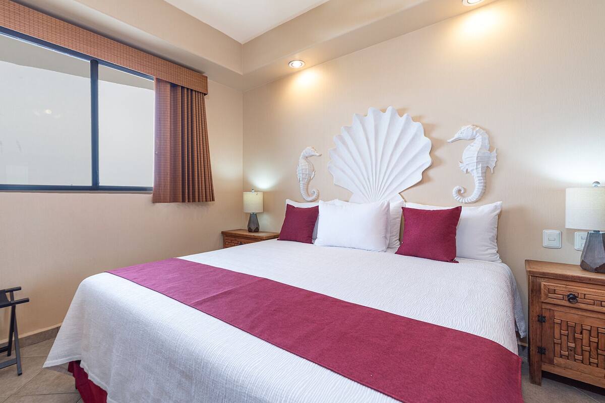 A hotel room with a large bed, a nightstand, a wooden cabinet, a window with curtains, and a headboard with decorative elements.