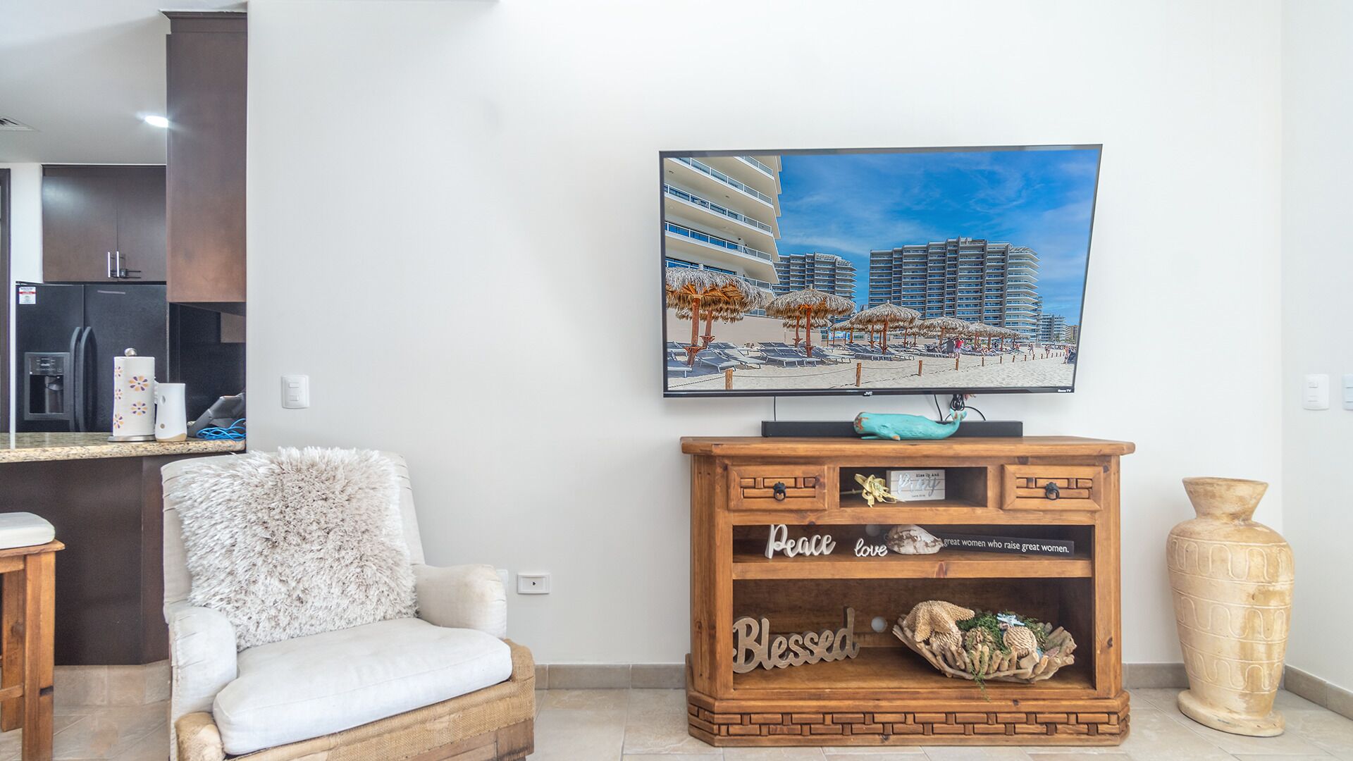 A modern living room with a flat-screen TV mounted on the wall, a wooden cabinet with decorative items, a fluffy chair, and a kitchen area in the background.