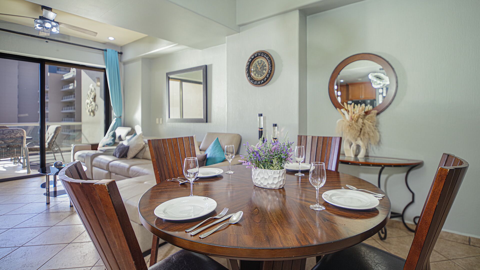 A dining area with a round wooden table set for four, surrounded by wooden chairs. There is a large mirror on the wall, a clock, and a decorative vase with flowers on the table.