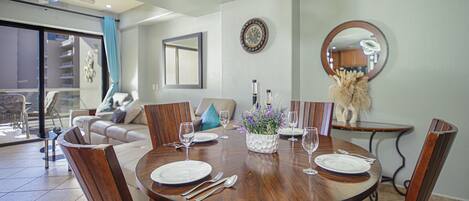 A dining area with a round wooden table set for four, surrounded by wooden chairs. There is a large mirror on the wall, a clock, and a decorative vase with flowers on the table.