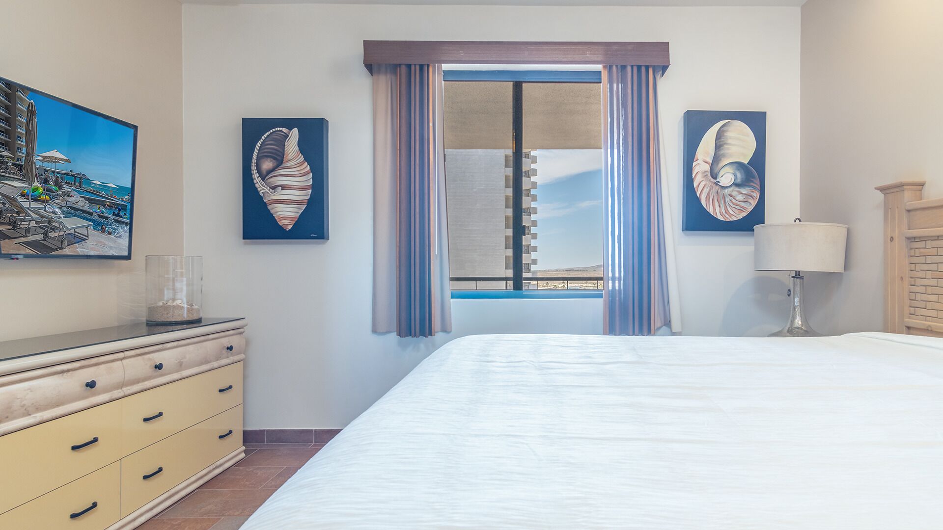 A hotel room with a bed, a dresser, a lamp, and two framed artworks on the wall.