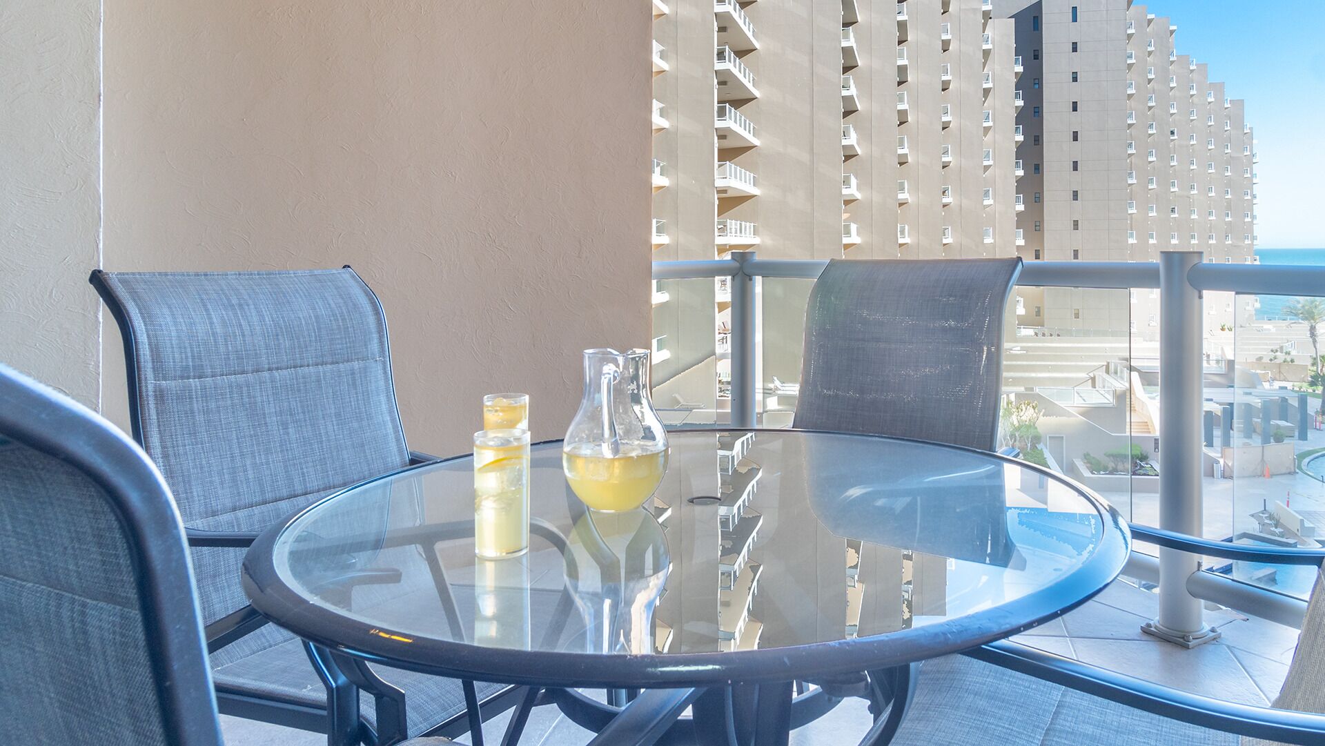 Un tavolo rotondo in vetro con due sedie, un boccale con una bevanda gialla e un bicchiere con una fetta di limone su un balcone che offre vista su un grattacielo.