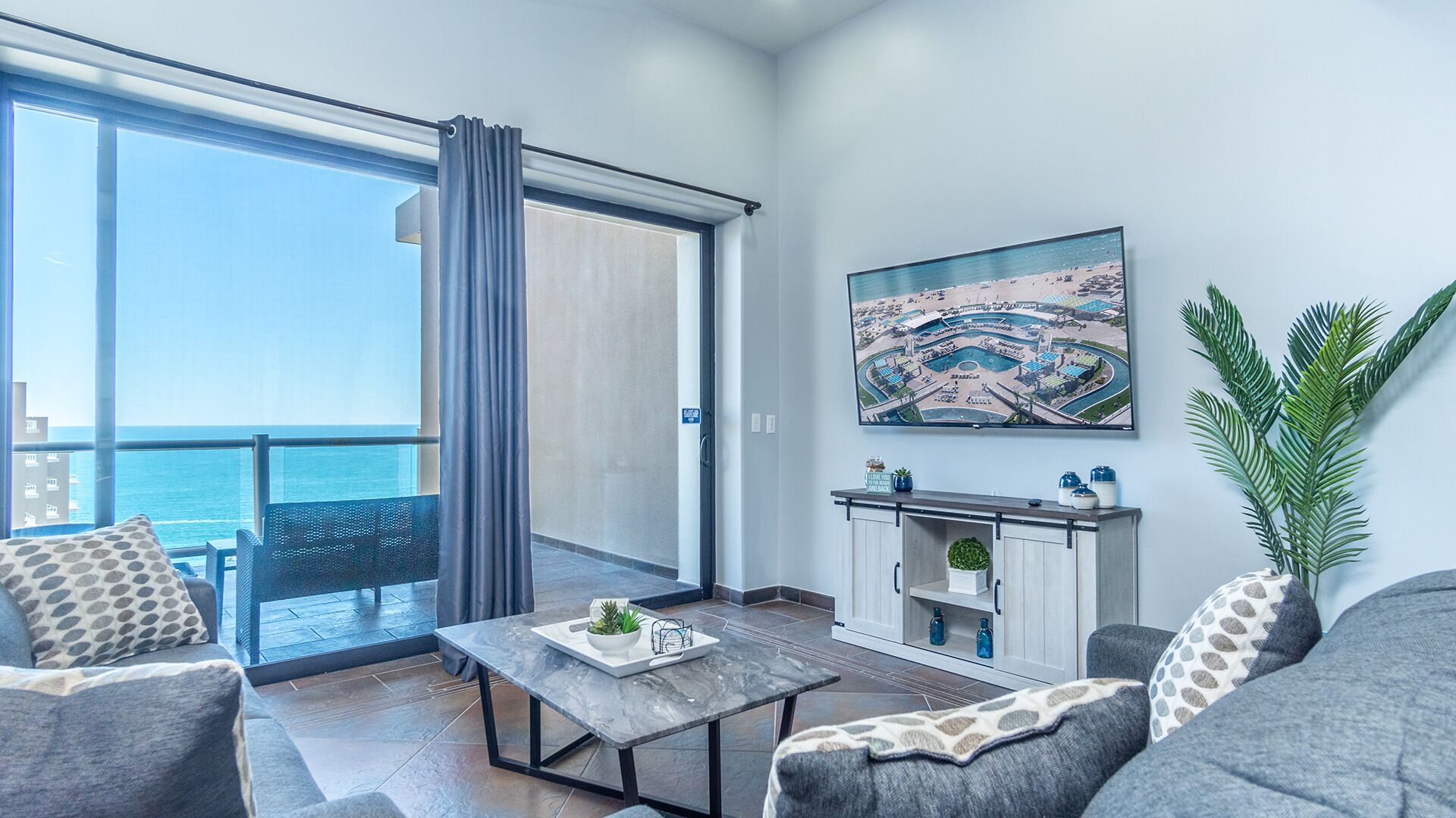 Ein modernes Wohnzimmer mit einem großen Schiebeglasfenster, das auf einen Balkon mit Meerblick führt. Es gibt ein Sofa, einen Couchtisch und eine Kommode mit Dekorationselementen.