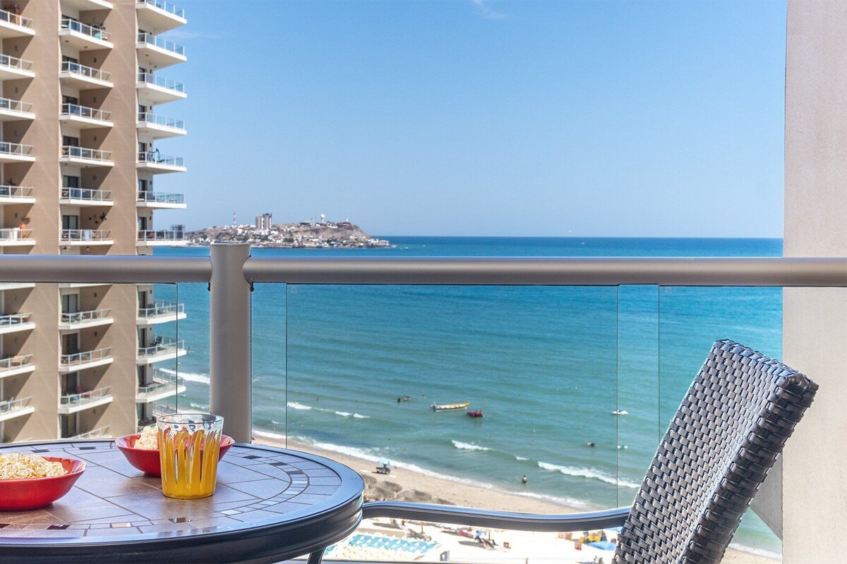 Un balcon avec une table dressée pour deux personnes, offrant une vue sur une plage et sur les bâtiments d’une ville lointaine.
