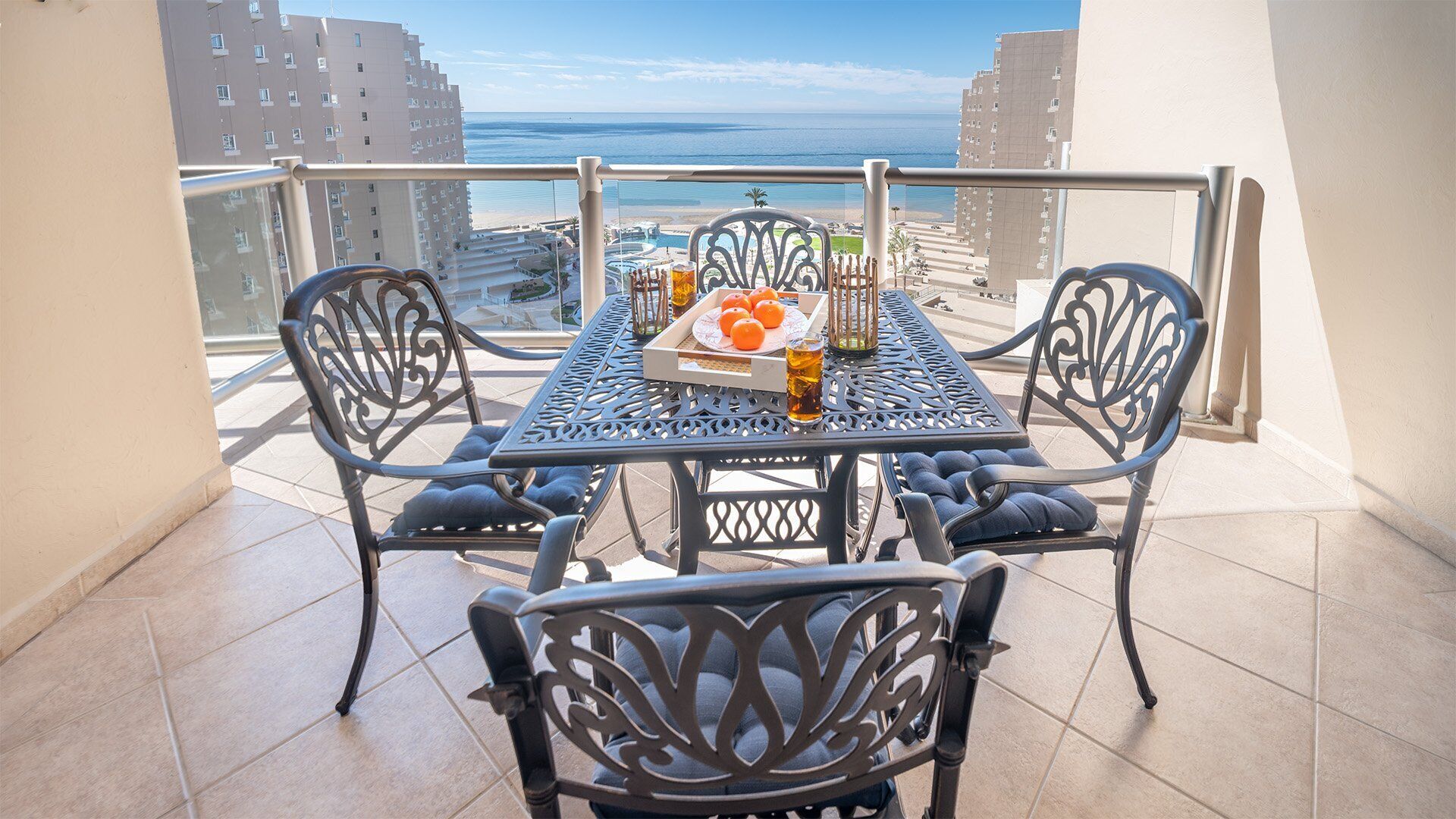 A balcony with a table set for two, featuring a tray with oranges and glasses, overlooking a beach and buildings.