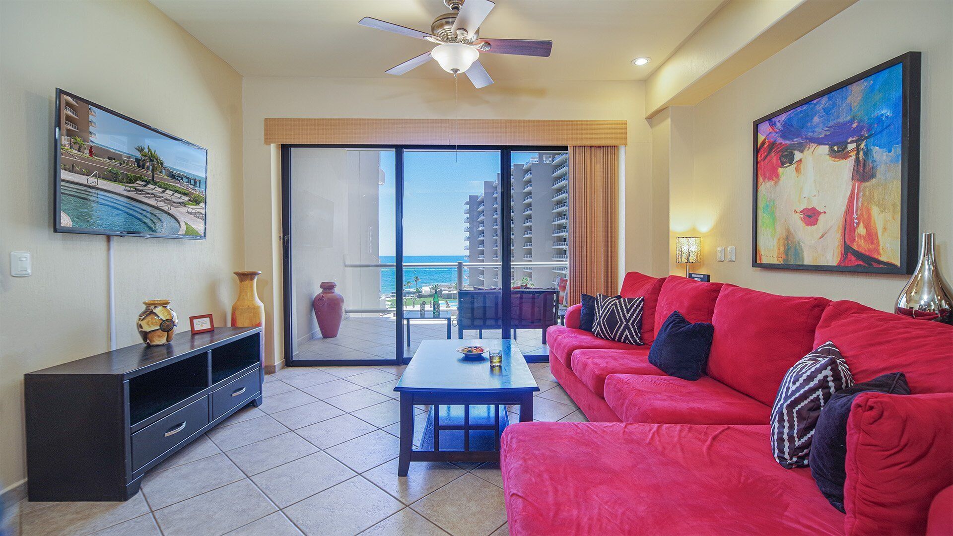A living room with a red sofa, a coffee table, and a TV on a stand. There is a balcony with a view of the ocean and buildings.