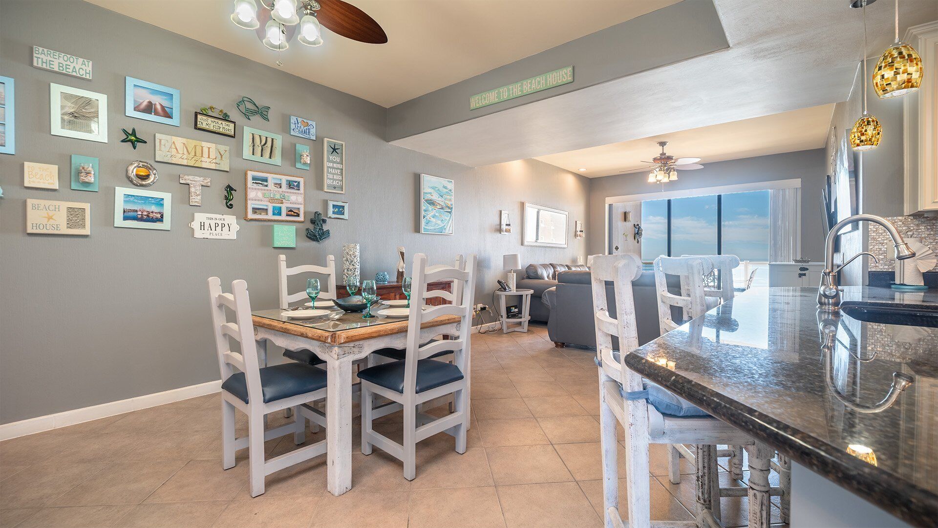 Una cocina moderna con comedor, que cuenta con una isla grande con pileta, sillas blancas y una mesa de comedor. Las paredes están decoradas con diversos adornos de temática playera.