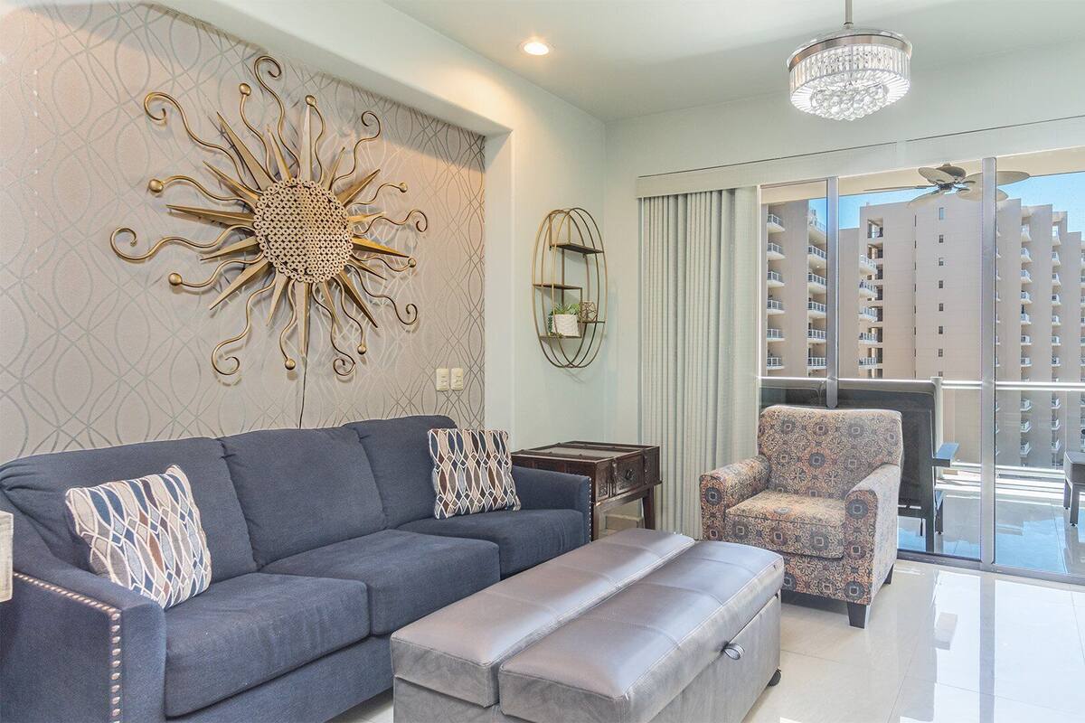 A modern living room with a grey sofa, patterned cushions, a decorative wall piece, a small side table, and a patterned armchair.